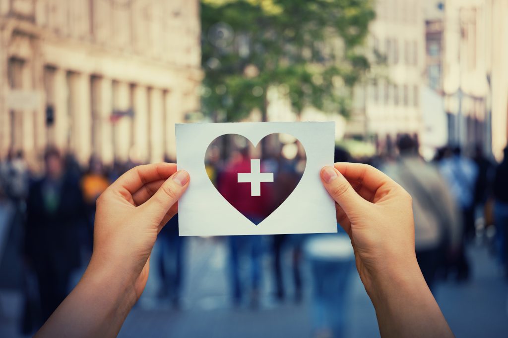 hands holding a paper sheet with heart and cross icon over a crowded street background.
